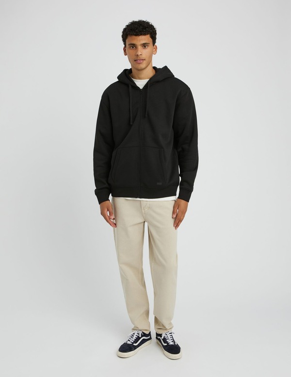 Sweat zippé noir porté ouvert sur un t-shirt blanc avec un chino beige et sneakers bicolores marine et blanc