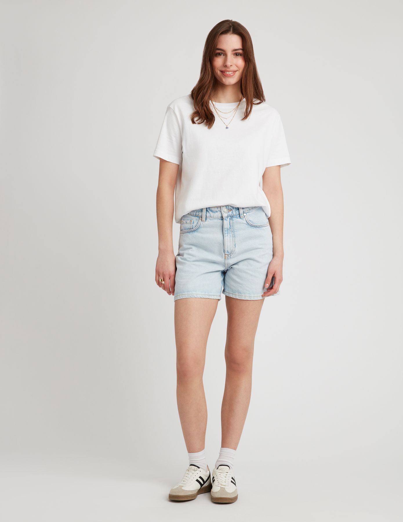 Femme en short mom fit en jean bleu clair délavé avec un t-shirt blanc oversize et des sneakers rétro blanches et noires