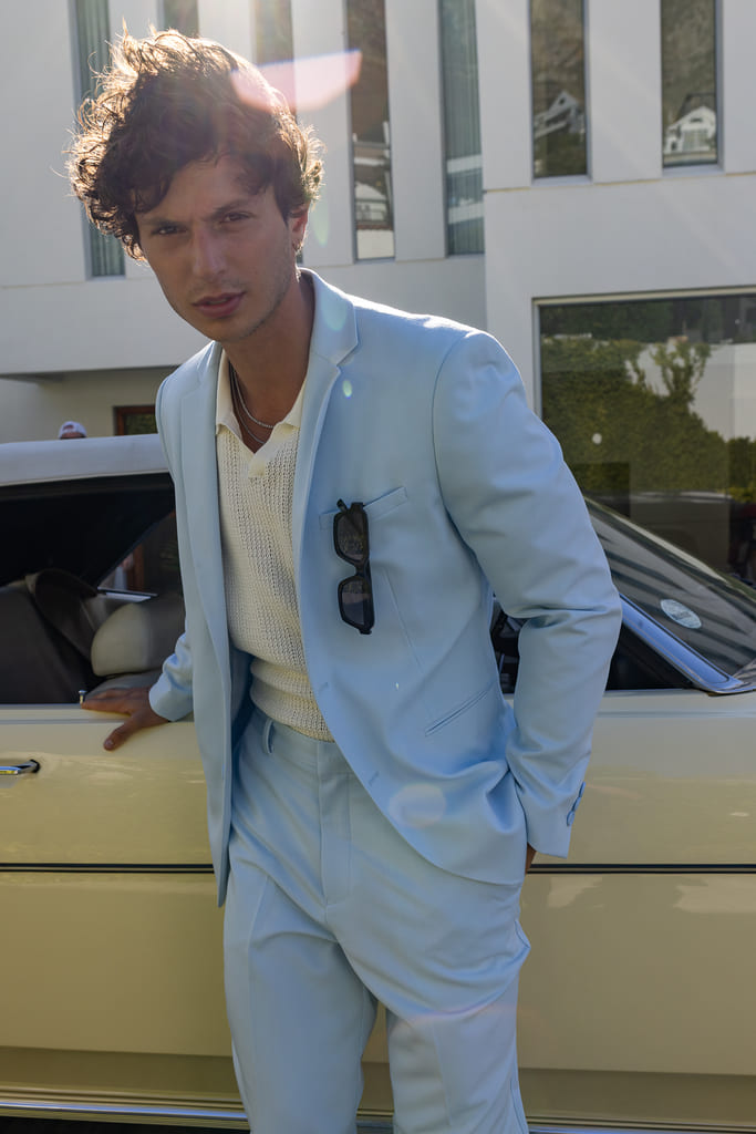 Costume bleu ciel pastel porté avec un pull col V en maille écrue et des lunettes de soleil glissées dans la poche poitrine