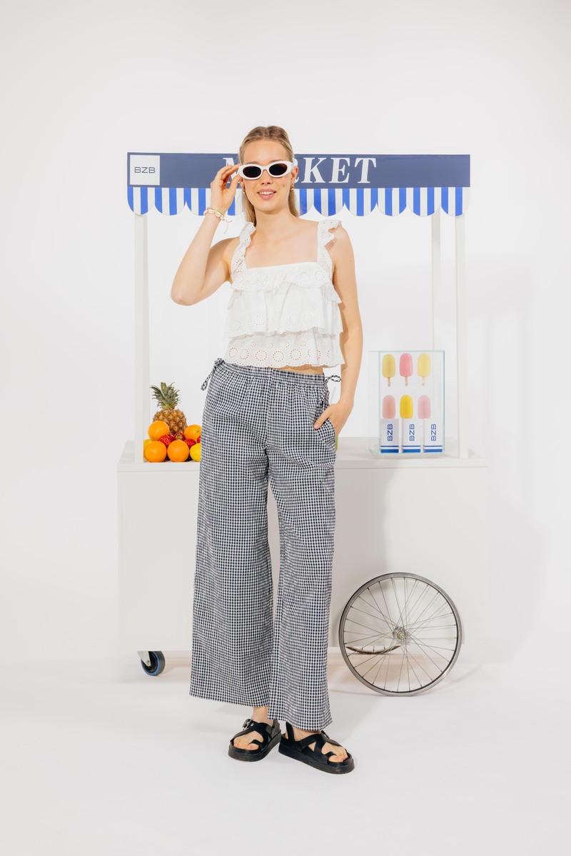 Combo tendance : crop top en broderie anglaise blanche à bretelles larges et volants étagés associé à un pantalon large vichy marine et blanc, avec sandales noires chunky et lunettes de soleil ovales blanches