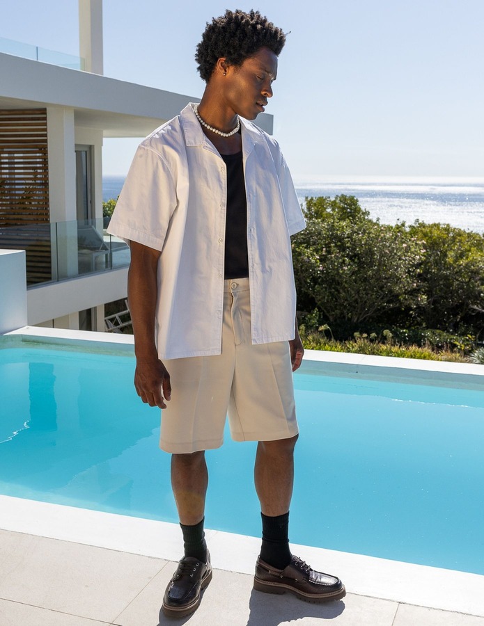 Look estival avec t-shirt noir, chemise blanche ouverte en layering, bermuda beige et mocassins chunky