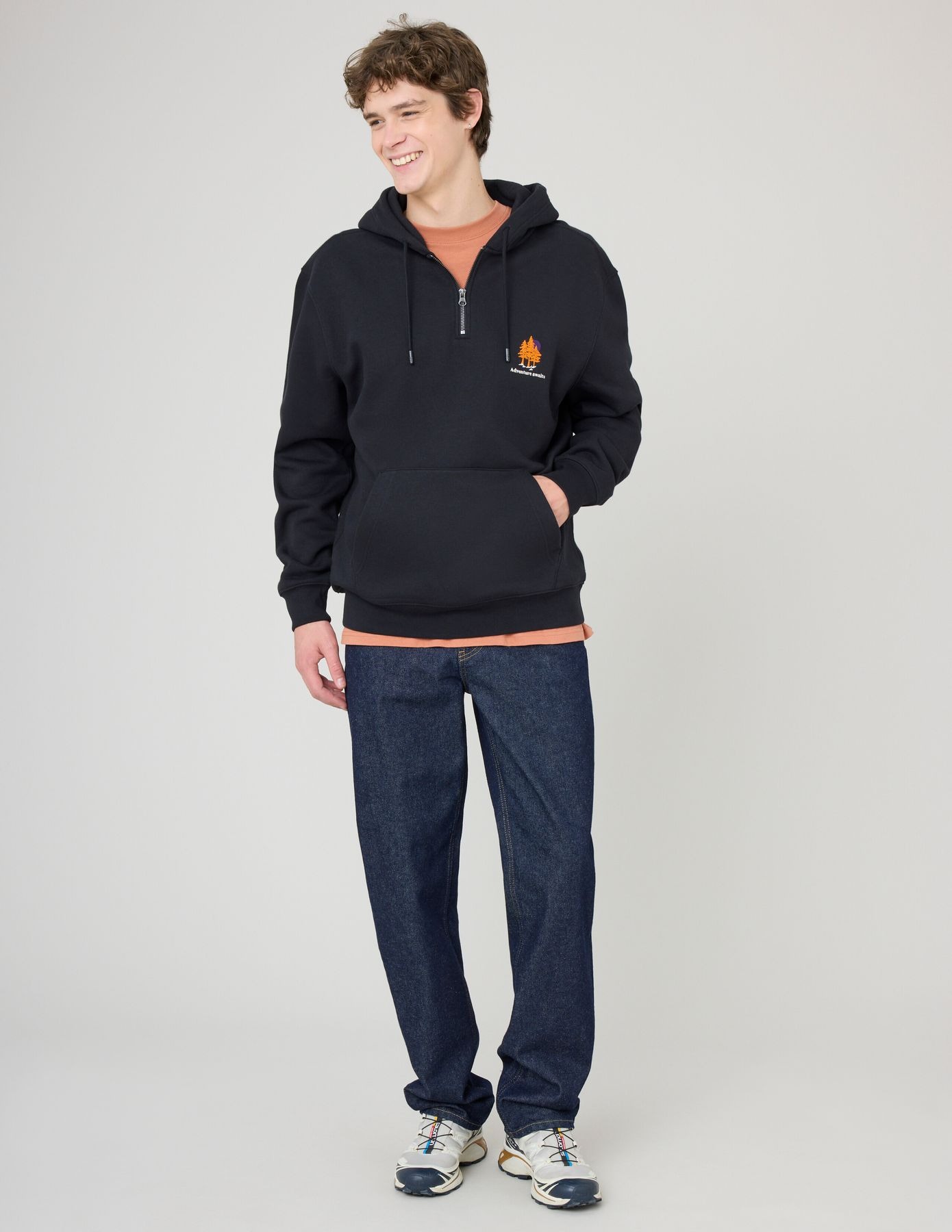 Homme en hoodie demi-zip noir brodé de sapins orangés avec un t-shirt terracotta et un jean brut foncé