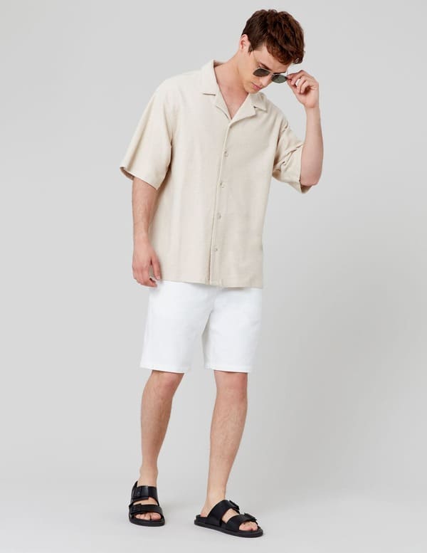 Homme en chemise camp collar beige sable oversize avec un bermuda blanc et des sandales noires