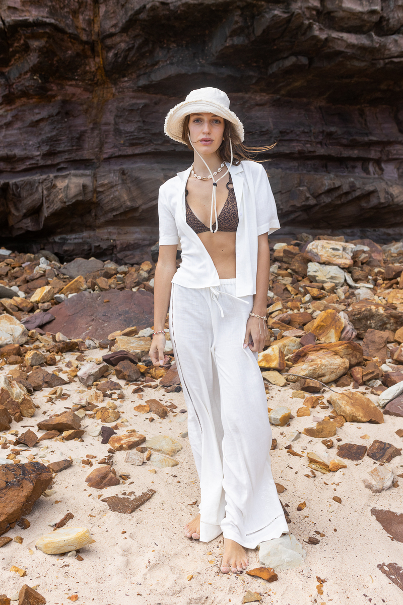 Look beach chic : chemise blanche à manches courtes portée ouverte sur un bikini léopard terracotta, pantalon large blanc à revers et bucket hat en tissu côtelé crème
