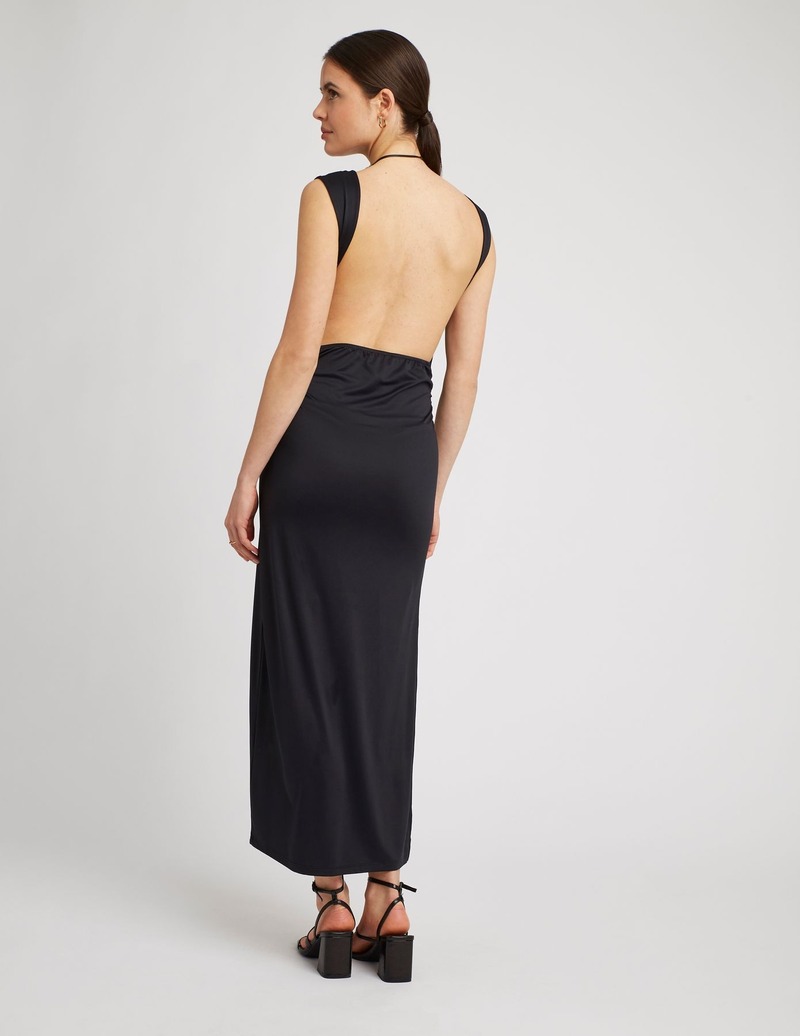Tenue de soirée estivale : robe longue noire dos nu intégralement dévoilé avec col halter noué, coupe droite jusqu'aux chevilles légèrement froncée sur les côtés, portée avec des sandales à lanières à talons carrés et une créole dorée