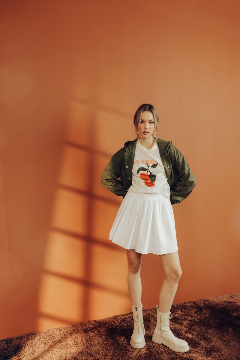 Look féminin décontracté : t-shirt graphique aux tons orangés, jupe patineuse blanche courte et bomber kaki porté ouvert sur les épaules, complété par des bottines zippées crème