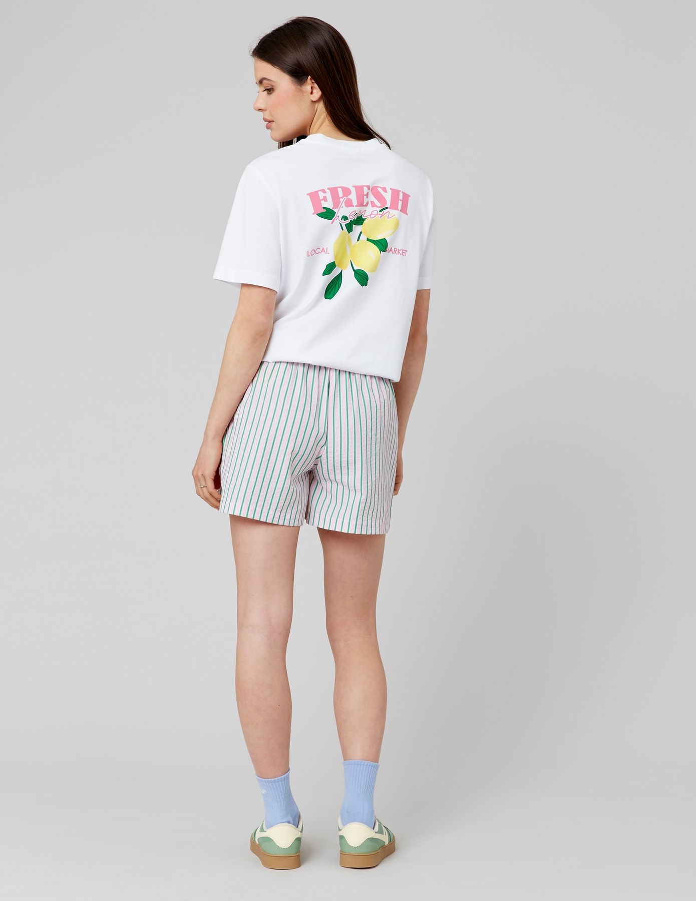 Look femme été coloré et joyeux avec un short à rayures roses et vertes ample associé à un t-shirt oversize blanc imprimé Fresh Lemon et des sneakers verts et crème