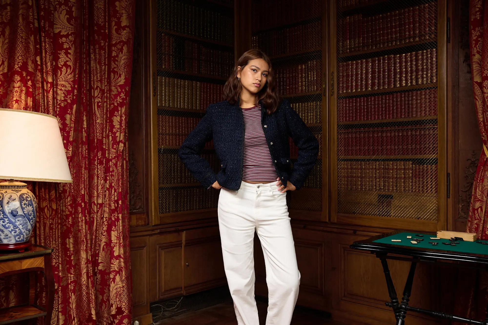 Veste tweed boutonnée portée sur un t-shirt rayé manches longues et un pantalon straight en velours, une loupe à la main, dans une bibliothèque ancienne