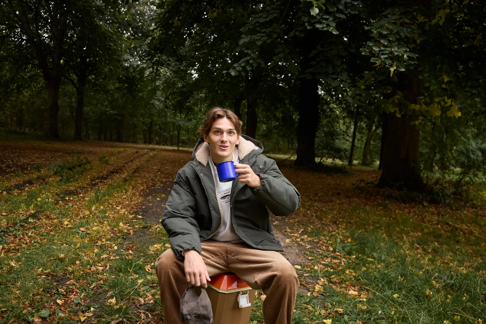 Parka à capuche portée sur un sweat brodé et un pantalon en velours côtelé, assis en forêt une tasse à la main