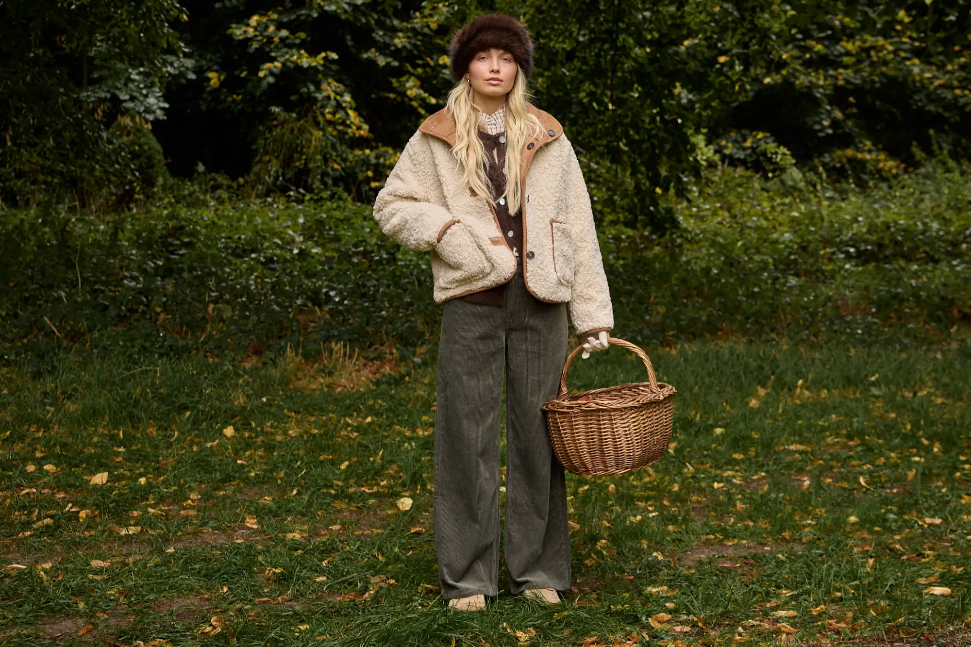Veste en sherpa réversible portée sur un gilet boutonné et un pantalon wide leg en velours, headband en fourrure, un panier à la main en forêt