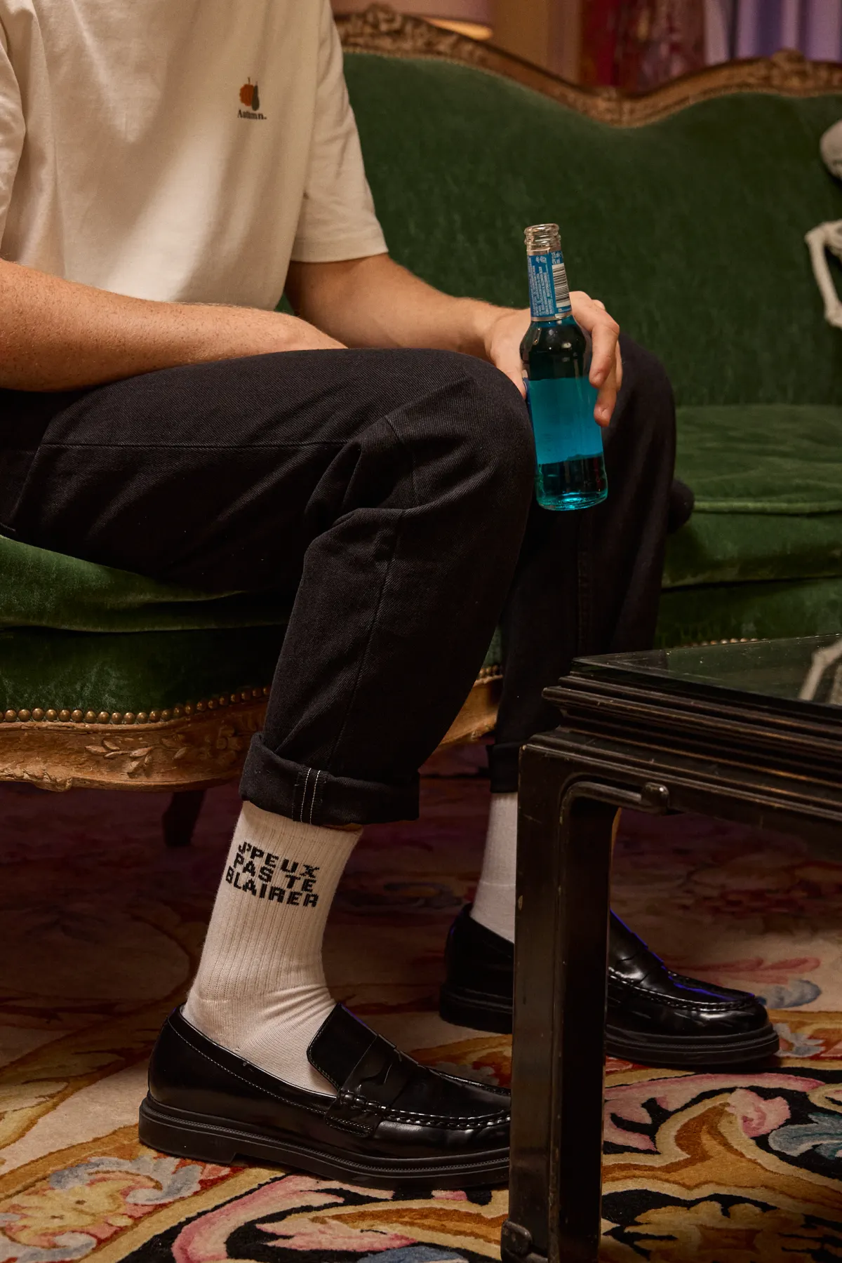 Mannequin assis sur un canapé de velours vert, en jean noir, mocassins et chaussettes blanches imprimées, une bouteille à la main