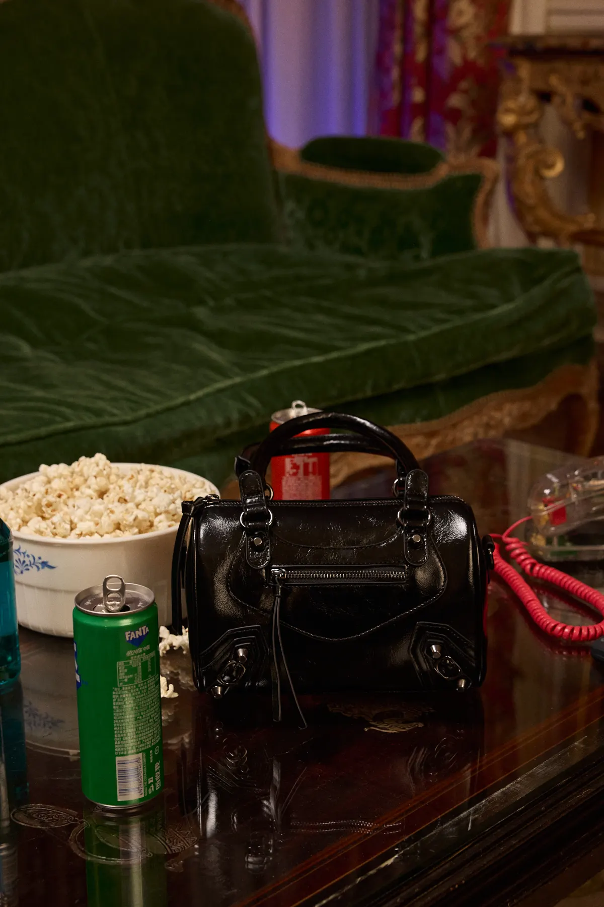 Sac à main bowling noir verni posé sur une table basse entre un pot de pop-corn et des canettes, dans un salon ancien
