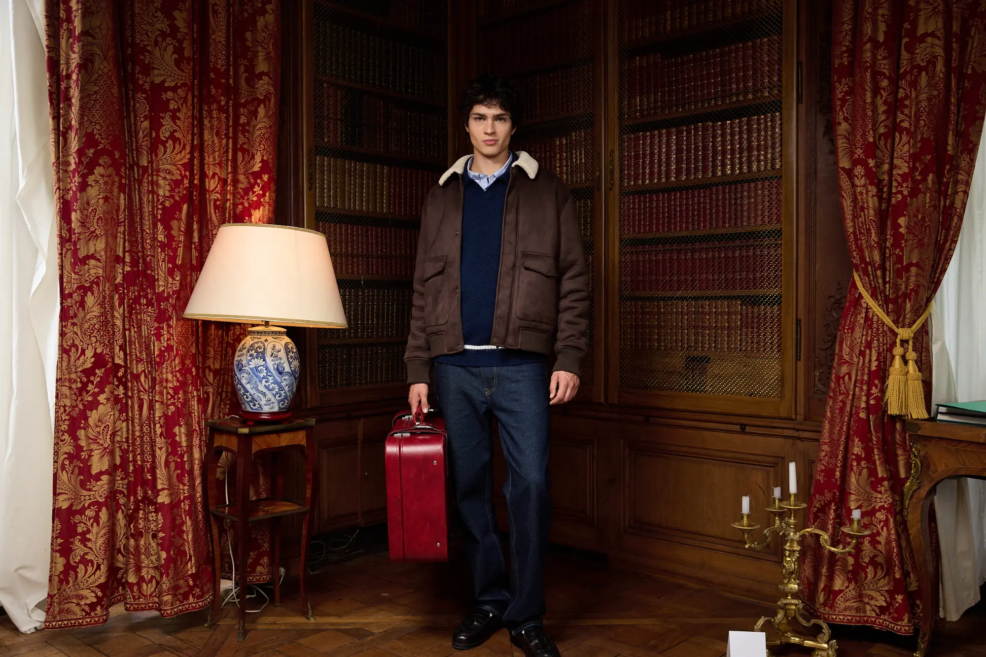 Blouson aviateur zippé porté sur un pull col polo liseret et un jean comfort brut, une valise rouge à la main, dans une bibliothèque ancienne