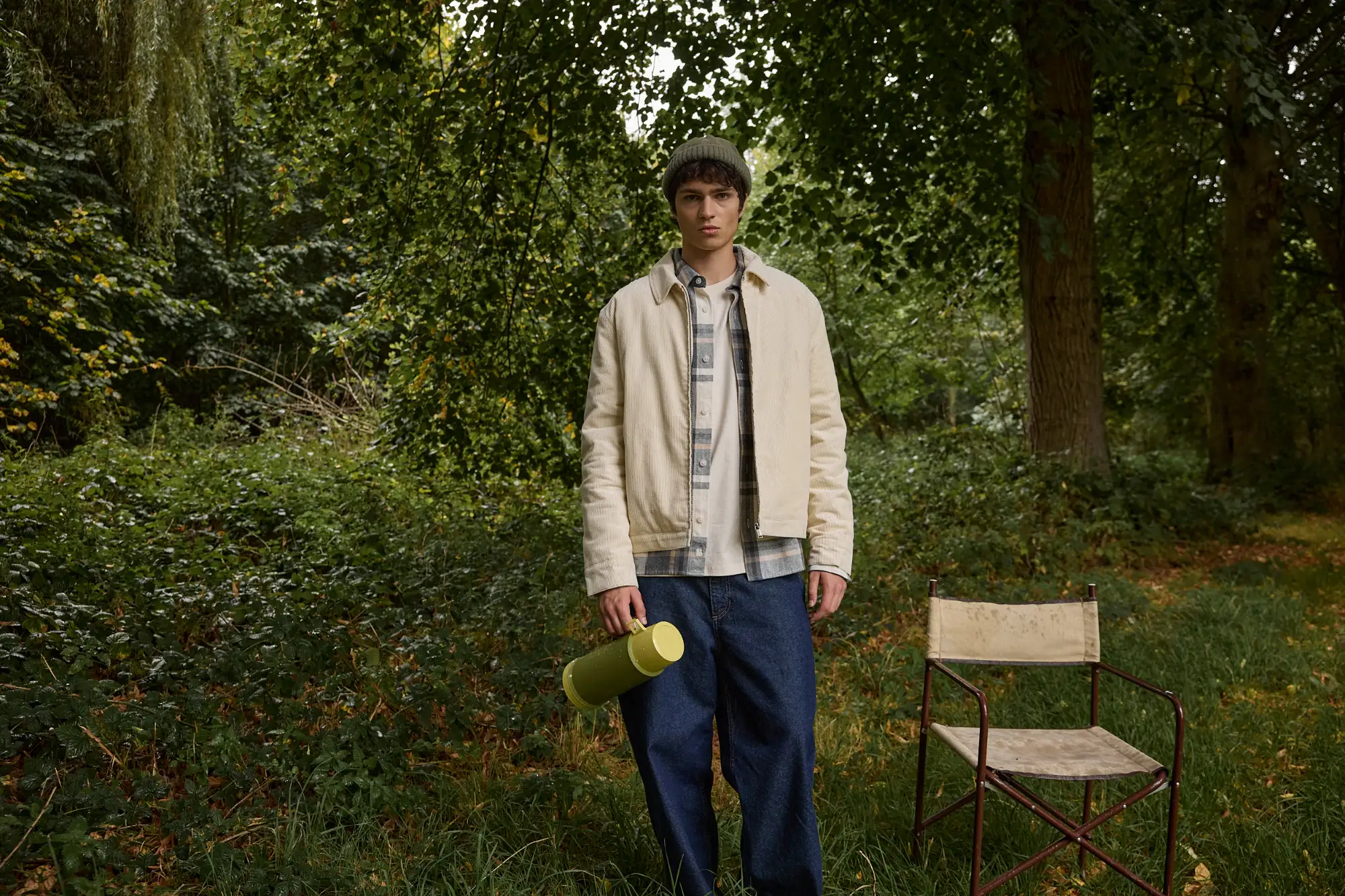 Veste velours zippée portée sur une chemise à carreaux et un jean skater brut, bonnet sur la tête et thermos à la main, en forêt